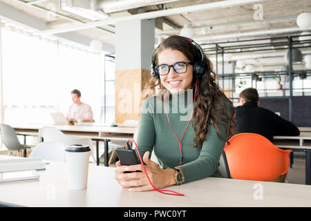 Fröhlich aufgeregt attraktiven weiblichen Manager in Brillen am Tisch mit takeout Kaffee saßen und an der Kamera, während Sie Musik hören über Smart Stockfoto