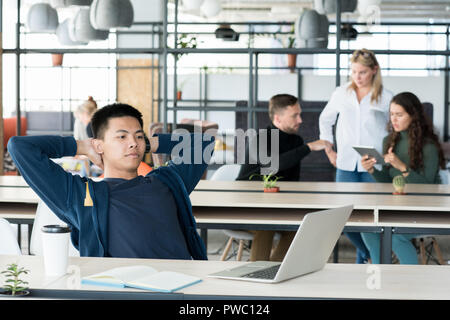 Ernsthafte entspannt Asiaten in Freizeitkleidung, Hände hinter dem Kopf und sitzen auf Stuhl beim Warten auf die Datei auf dem Laptop zu laden. Stockfoto