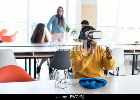 Ernsthafte neugierige junge Mitte - östliche hipster Mann mit Bart am Tisch sitzen und halten unsichtbares Objekt beim Spielen video game in VR-Headset Stockfoto