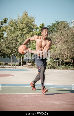 Eine afroamerican junger Mann ohne T-Shirt ist Basketball spielen in einem Park in Madrid im Sommer mittags. Er macht einen super Sprung eine Dun zu machen Stockfoto