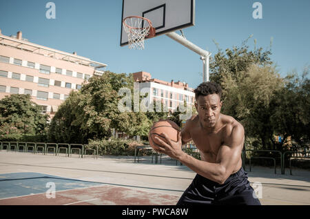 Nahaufnahme einer afroamerican junger Mann ohne T-Shirt ist Basketball spielen in einem Park in Madrid im Sommer mittags. Er sucht an der Kamera Stockfoto