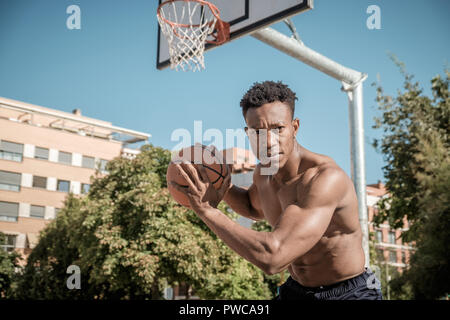 Nahaufnahme einer afroamerican junger Mann ohne T-Shirt ist Basketball spielen in einem Park in Madrid im Sommer mittags. Er sucht an der Kamera Stockfoto