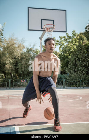 Eine afroamerican junger Mann ohne T-Shirt ist Basketball spielen in einem Park in Madrid im Sommer mittags. Er springt die Kugel unter dem Korb. Stockfoto