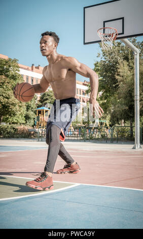 Eine afroamerican junger Mann ohne T-Shirt ist Basketball spielen in einem Park in Madrid im Sommer mittags. Er springt die Kugel unter dem Korb. Stockfoto