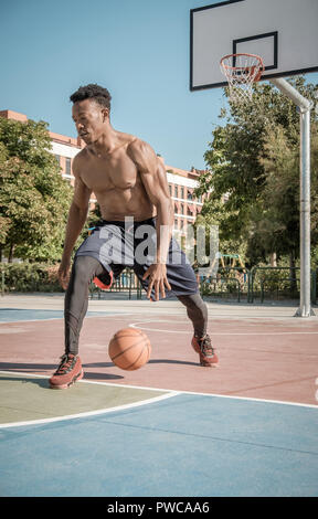 Eine afroamerican junger Mann ohne T-Shirt ist Basketball spielen in einem Park in Madrid im Sommer mittags. Er springt die Kugel unter dem Korb. Stockfoto