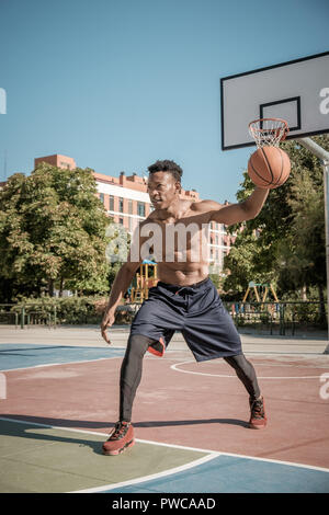 Eine afroamerican junger Mann ohne T-Shirt ist Basketball spielen in einem Park in Madrid im Sommer mittags. Er springt die Kugel unter dem Korb. Stockfoto