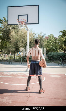 Eine afroamerican junger Mann ohne T-Shirt ist Basketball spielen in einem Park in Madrid im Sommer mittags. Er steht mit dem Ball auf der einen Seite Stockfoto