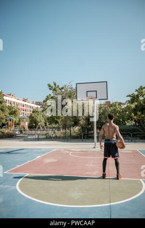 Eine afroamerican junger Mann ohne T-Shirt ist Basketball spielen in einem Park in Madrid im Sommer mittags. Er steht mit dem Ball auf der einen Seite Stockfoto