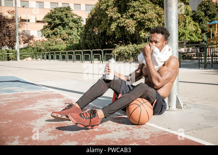 Eine afroamerican junger Mann ohne T-Shirt ist Basketball spielen in einem Park in Madrid im Sommer mittags. Er ist auf dem Gericht sitzt und Trinken Stockfoto