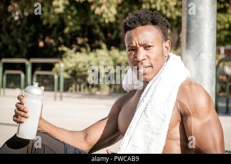 Eine afroamerican junger Mann ohne T-Shirt ist Basketball spielen in einem Park in Madrid im Sommer mittags. Er ist auf dem Gericht sitzt und Trinken Stockfoto