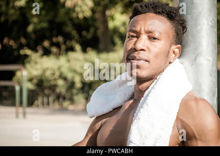 Eine afroamerican junger Mann ohne T-Shirt ist Basketball spielen in einem Park in Madrid im Sommer mittags. Er ist auf dem Gericht sitzt und Trinken Stockfoto