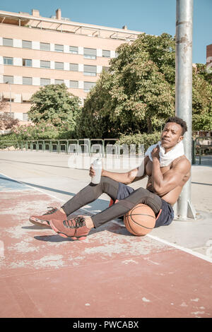 Eine afroamerican junger Mann ohne T-Shirt ist Basketball spielen in einem Park in Madrid im Sommer mittags. Er ist auf dem Gericht sitzt und Trinken Stockfoto