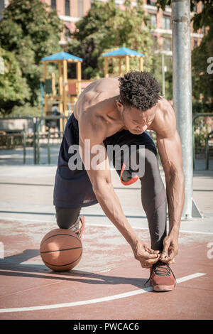 Eine afroamerican junger Mann ohne T-Shirt ist Basketball spielen in einem Park in Madrid im Sommer mittags. Er ist auf seine Knie, seine Schuhe binden mit t Stockfoto