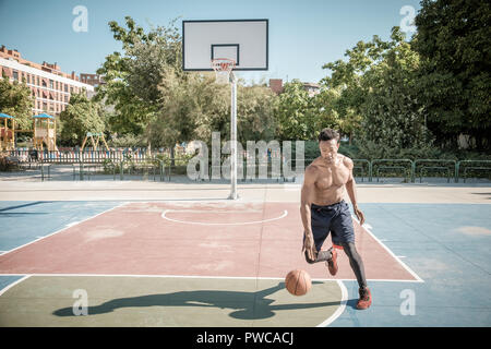 Eine afroamerican junger Mann ohne T-Shirt ist Basketball spielen in einem Park in Madrid im Sommer mittags. Er springt die Kugel unter dem Korb. Stockfoto