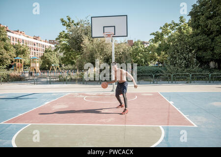 Eine afroamerican junger Mann ohne T-Shirt ist Basketball spielen in einem Park in Madrid im Sommer mittags. Er springt die Kugel unter dem Korb. Stockfoto