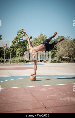 Eine afroamerican junger Mann ohne T-Shirt ist das Gleichgewicht mit einem Basketball in einem Park in Madrid im Sommer mittags. Stockfoto