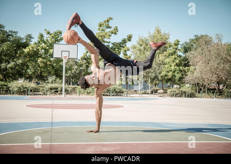 Eine afroamerican junger Mann ohne T-Shirt ist das Gleichgewicht mit einem Basketball in einem Park in Madrid im Sommer mittags. Stockfoto