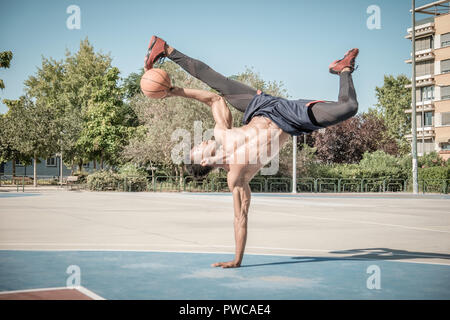 Eine afroamerican junger Mann ohne T-Shirt ist das Gleichgewicht mit einem Basketball in einem Park in Madrid im Sommer mittags. Stockfoto