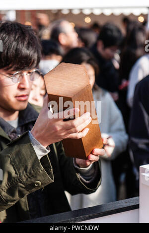 Japanische neues Jahr, shogatsu. Nahaufnahme der männlichen Händen hält Omikuji, während andererseits Schlepper Papierstreifen durch die Öffnung im Deckel. Ikuta Schrein. Stockfoto