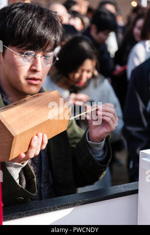 Japanische neues Jahr, shogatsu. Nahaufnahme der männlichen Händen hält Omikuji, während andererseits Schlepper Papierstreifen durch die Öffnung im Deckel. Ikuta Schrein. Stockfoto