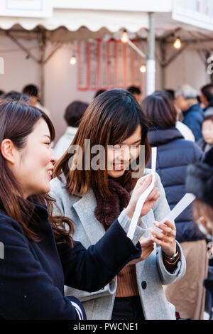 Japanische neues Jahr, shogatsu. Nahaufnahme, zwei junge Frauen, kichern, während sie ihre Omikuji Papier Vermögen lesen Sie rutscht bei der shinto Ikuta Heiligtum in Kobe. Stockfoto