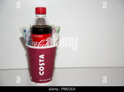 Costa Kaffee Tasse mit Coca Cola Flasche innen mit Bargeld auf jeder Seite auf weißem Hintergrund. Stockfoto