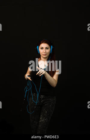 Auf schwarzem Hintergrund, nette junge Frau mit langen roten Haaren trägt blaue Kopfhörer, sie ist glücklich, sie tanzt und singt Stockfoto