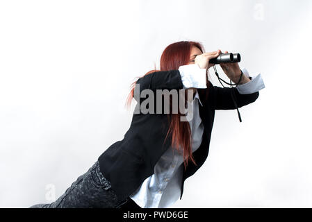 Eine Frau mit einem Fernglas in der Hand auf einem hellen Hintergrund steht, ist sie in einer schwarzen Jacke und weissen Hemd bekleidet, ist sie auf der Suche nach Etwas Stockfoto