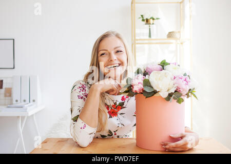 Gerne blonde Frau in einem modernen Büro mit Blumen in einen Hut. Strauß Pfingstrosen. Blume Lieferung an den Arbeitsplatz Stockfoto