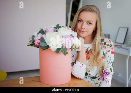 Gerne blonde Frau in einem modernen Büro mit Blumen in einen Hut. Strauß Pfingstrosen. Blume Lieferung an den Arbeitsplatz Stockfoto