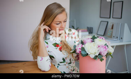 Gerne blonde Frau in einem modernen Büro mit Blumen in einen Hut. Strauß Pfingstrosen. Blume Lieferung an den Arbeitsplatz Stockfoto