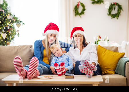 Ein kleiner Junge mit Mutter und Großmutter mit Santa Hut auf einem Sofa zu Hause sitzen zur Weihnachtszeit heraus haften Zungen. Stockfoto