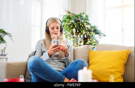 Eine junge Frau mit Kopfhörern und Smartphone sitzt auf einem Sofa zu Hause an Weihnachten, Musik hören. Stockfoto