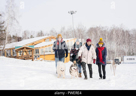 In voller Länge Porträt von zwei junge Paare genießen die Zeit im Skigebiet zu Fuß Hunde draußen und glücklich lächelnd zu Fuß in Richtung Kamera gegen Winter Stockfoto