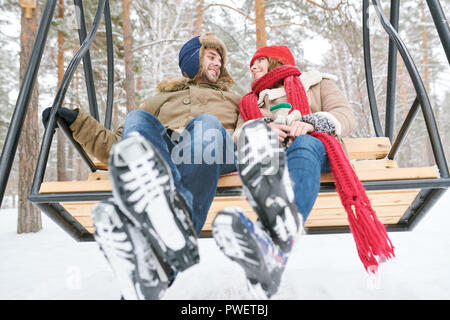 Volle Länge niedrigen Winkel Portrait von glückliches junges Paar auf hölzernen schwingen und sich gegenseitig zärtlich, während Datum genießen im Winter Wald Stockfoto
