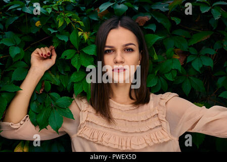 Junge Frau lehnt sich auf einem Stein Zaun mit grünen Blättern und Suchen in die Kamera. Stockfoto