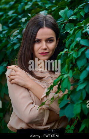 Portrait einer jungen selbstbewusste Frau lehnte sich auf einem Stein Zaun mit grünen Blättern und Suchen in die Kamera. Stockfoto