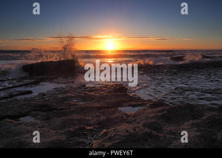 Sonnenuntergang mit orange Wolken und Spray von Wellen auf die Felsen an der Dunraven Bay, Tal von Glamorgan, South Wales Stockfoto