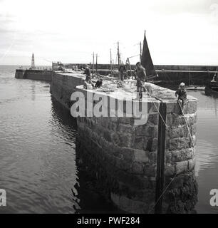 1950, historische, junge Kinder Angeln vom Meer an der Wand, in einem Hafen in Irland, im Vereinigten Königreich. In dieser Zeit, die Kinder hatten viel Freiheit außerhalb auf Ihren Selbst zu spielen und, wie in diesem Bild zu sehen sind, eine Gruppe von Jungen werden gerne Angeln von einer hohen Mauer ins tiefe Wasser unten ohne Aufsicht von Erwachsenen. Stockfoto