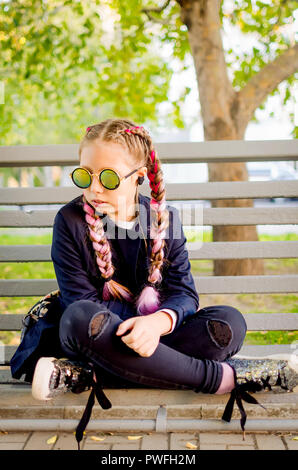 Portrait blond teen Mädchen mit rosa Kanekalon im Haar und einem Spiegel Sonnenbrille, sitzt auf einer Bank mit Beinen im Herbst Park in sonniger Tag verdreht Stockfoto