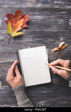 Mockup mit weißem Papier Notebook eingerichtet Herbst Blätter auf Holztisch. Woman's Hand schreiben im Editor ein. Ansicht von oben, senkrecht.