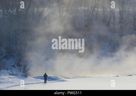 Die Silhouette eines Mannes, der an der Küste des zugefrorenen Fluss mit Nebel leuchtet durch die Morgensonne im Winter Stockfoto