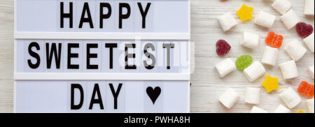 Lightbox mit Text "Happy süssesten Tag' und Süßigkeiten auf weißem Holz- Hintergrund, Ansicht von oben. Flach, von oben. Stockfoto