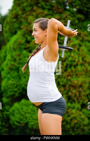 Zuversichtlich werdende Mutter tun overhead Trizeps Erweiterungen mit Hantel in Park Stockfoto