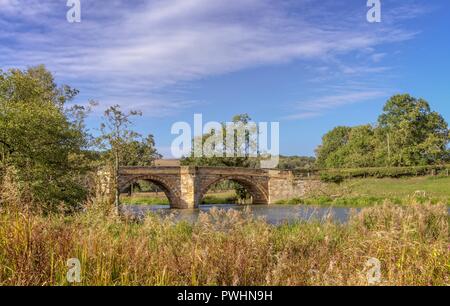 Die Bögen aus dem 19. Jahrhundert Brücke überspannt den Fluss Derwent. Gras Banken sind auf beiden Seiten und ein blauer Himmel mit Wolken ist oben. Stockfoto