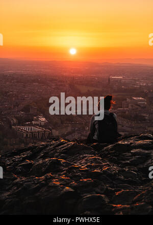 Das Mädchen von der Rückseite auf einem Felsen, der einen hohen Berg sitzt den Sonnenuntergang über der Stadt, Edinburgh, Scontald, Vereinigtes Königreich Stockfoto