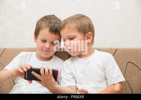 Zwei kleine Jungen in den weißen T-Shirts auf dem Sofa zu Hause sitzen und Smartphone zusammen anschauen Stockfoto