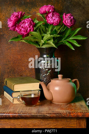 Schönen lila Päonien in eine Vase, eine Tasse Tee, ein Wasserkocher und Bücher auf einem Rost Hintergrund. Stockfoto