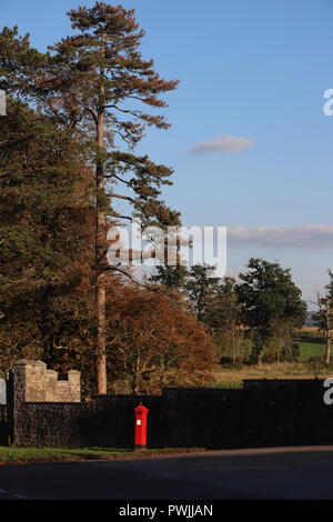 Die Sonne scheint auf eine helle rote Britische Post Box stand vor einer dunklen Mauer aus Stein, mit bepflanzten Felder und englischen Landschaft im Hintergrund. Stockfoto