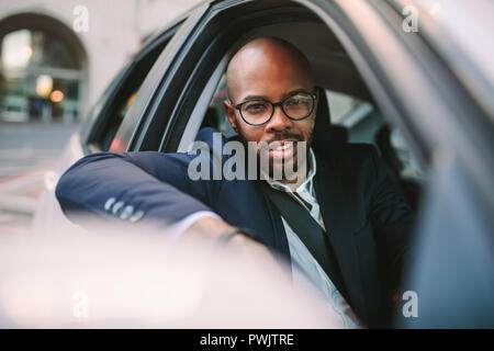 Afrikanischen Geschäftsmann in Anzug peeking aus dem Autofenster, und wenn man die Kamera während der Fahrt. Gut aussehender Geschäftsmann in Anzug fahren Auto auf die City Road. Stockfoto